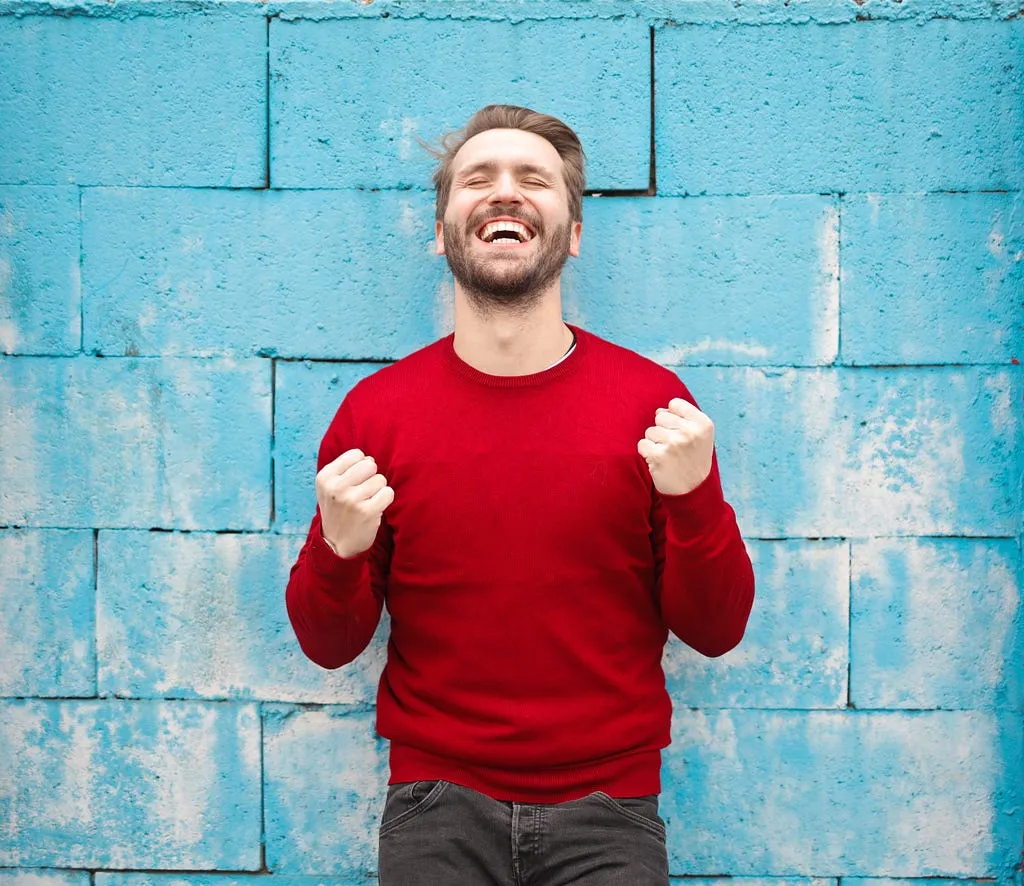 A man in a red jumper cheering with both fists clenched, in front of a blue wall